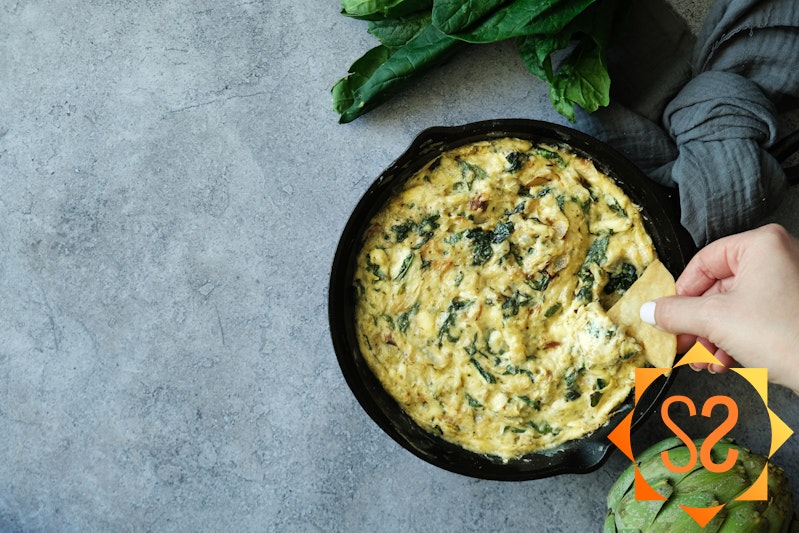 a hand dipping a chip into vegan spinach and artichoke dip