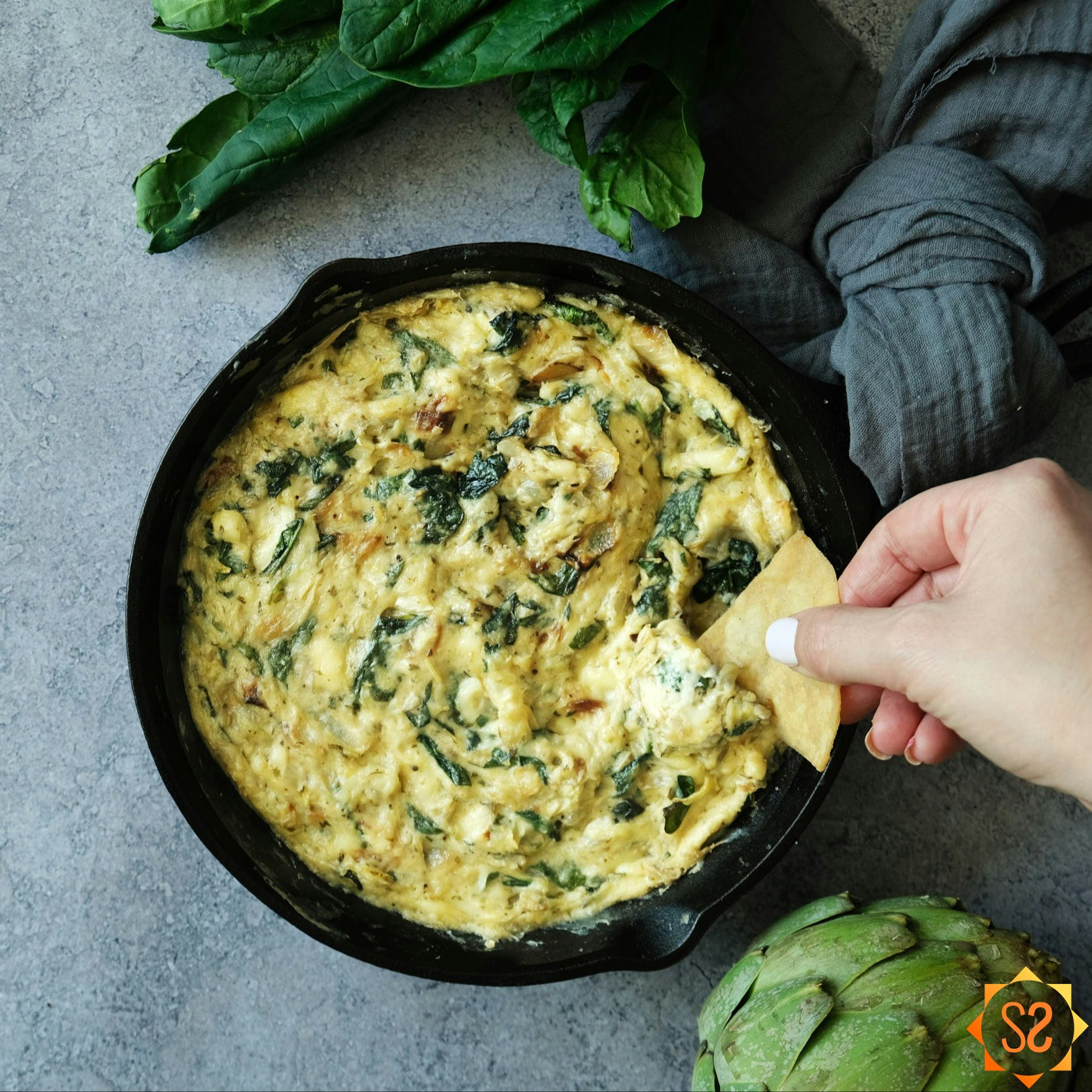 A hand dipping a chip into vegan spinach and artichoke dip.