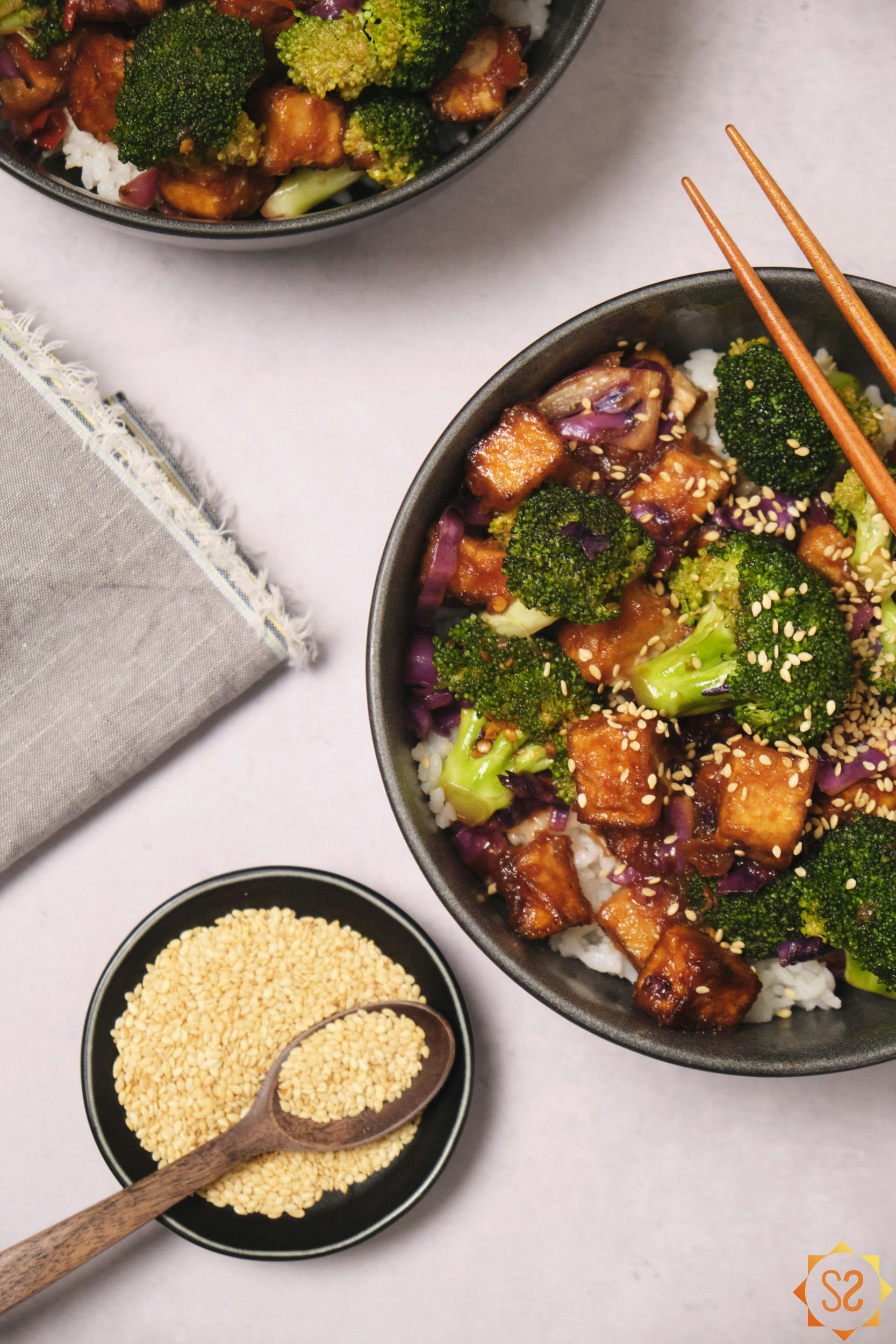 Two bowls of tofu stir-fry on a table with a small pinch bowl of sesame seeds on the side. 