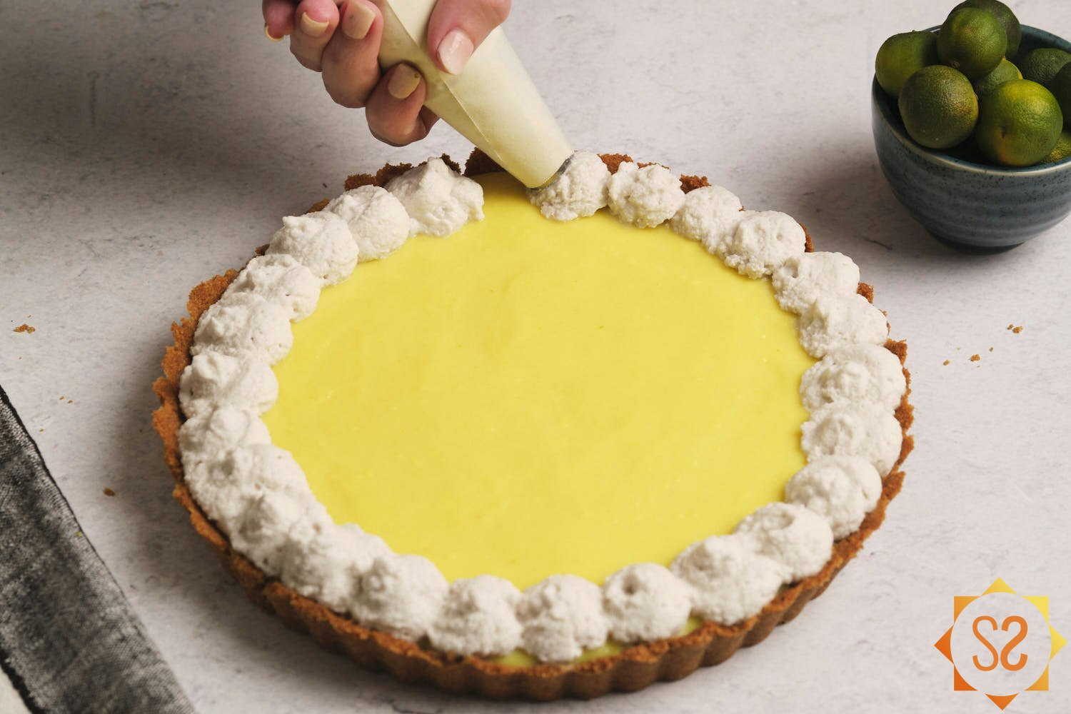 An image of hands piping vegan whipped cream onto a finished vegan Key lime pie, with a bowl of Key limes to the side.