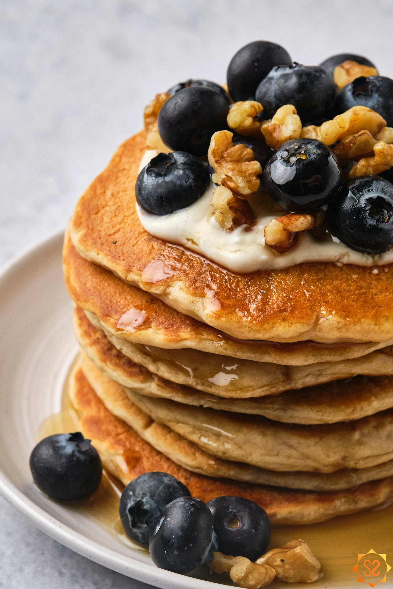 A close up view of a stack of vegan protein pancakes showing the toppings: cashew cream, blueberries, walnuts, maple syrup.