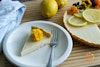 A slice of lemon tart on a plate with the whole tart and lemons to the side.
