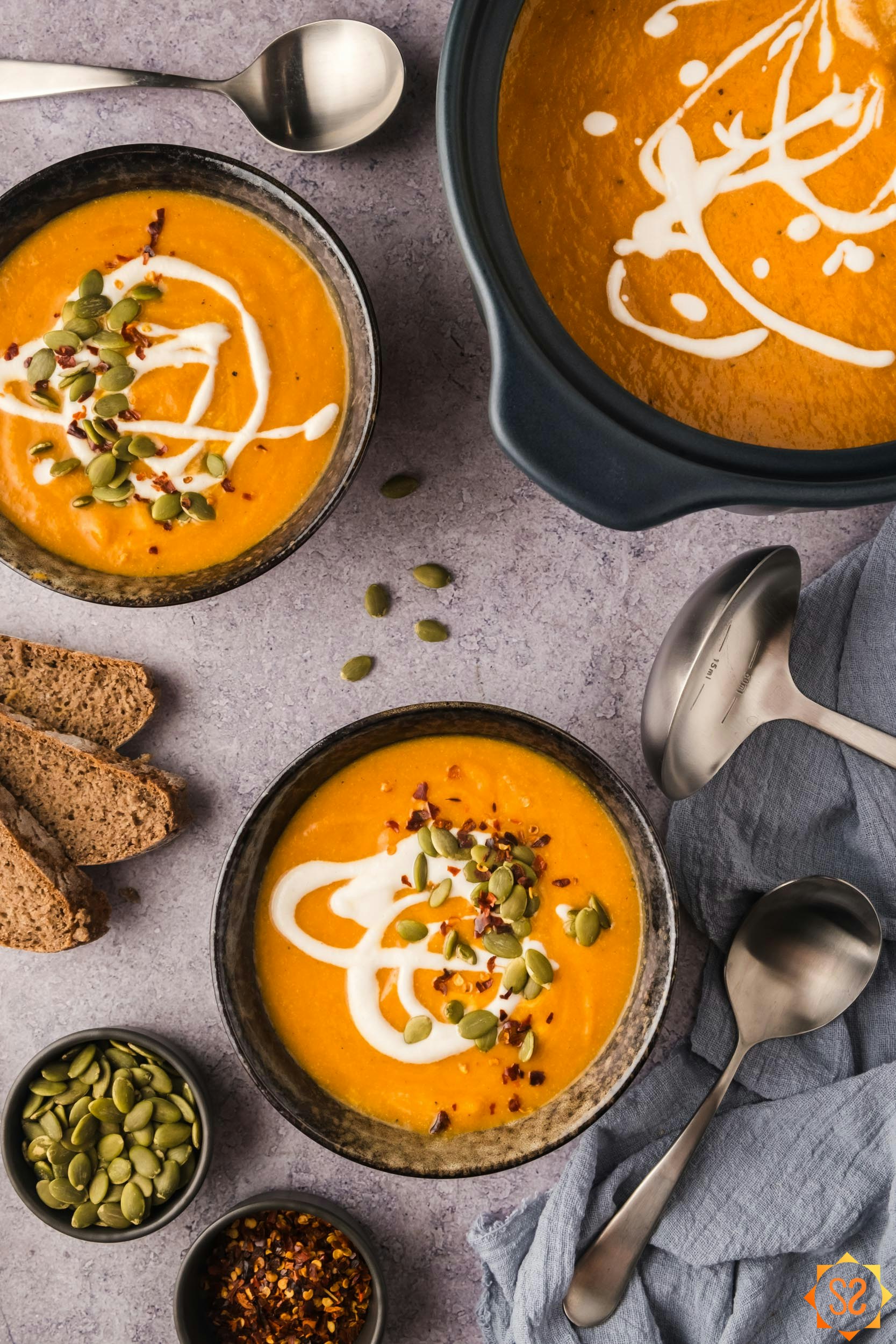 A top-down view of a table with two bowls and a pot of carrot pumpkin soup, along with spoons, bread, seeds, and red pepper.