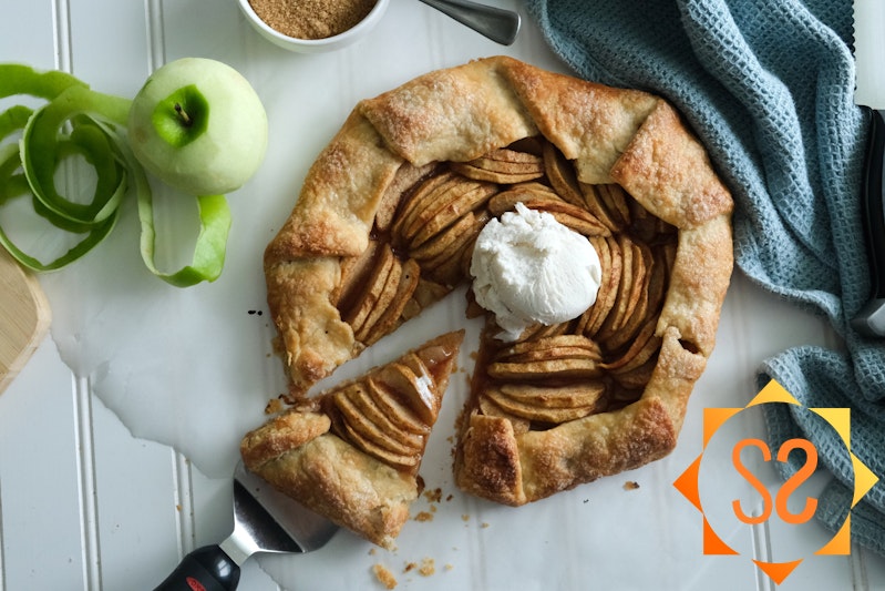 An apple galette with a slice being pulled away, topped with vegan ice cream; a peeled apple to the left, and a ramekin of brown sugar above.