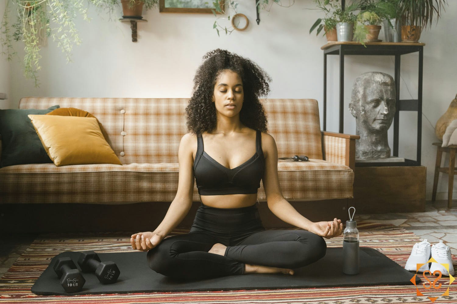 A woman meditating on a yoga mat in her living room, with weights and a water bottle to her sides.