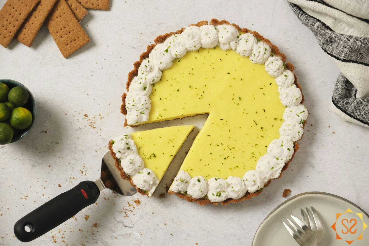 A vegan Key lime pie with a slice being removed, surrounded by graham crackers, a bowl of Key limes, and plates and forks.