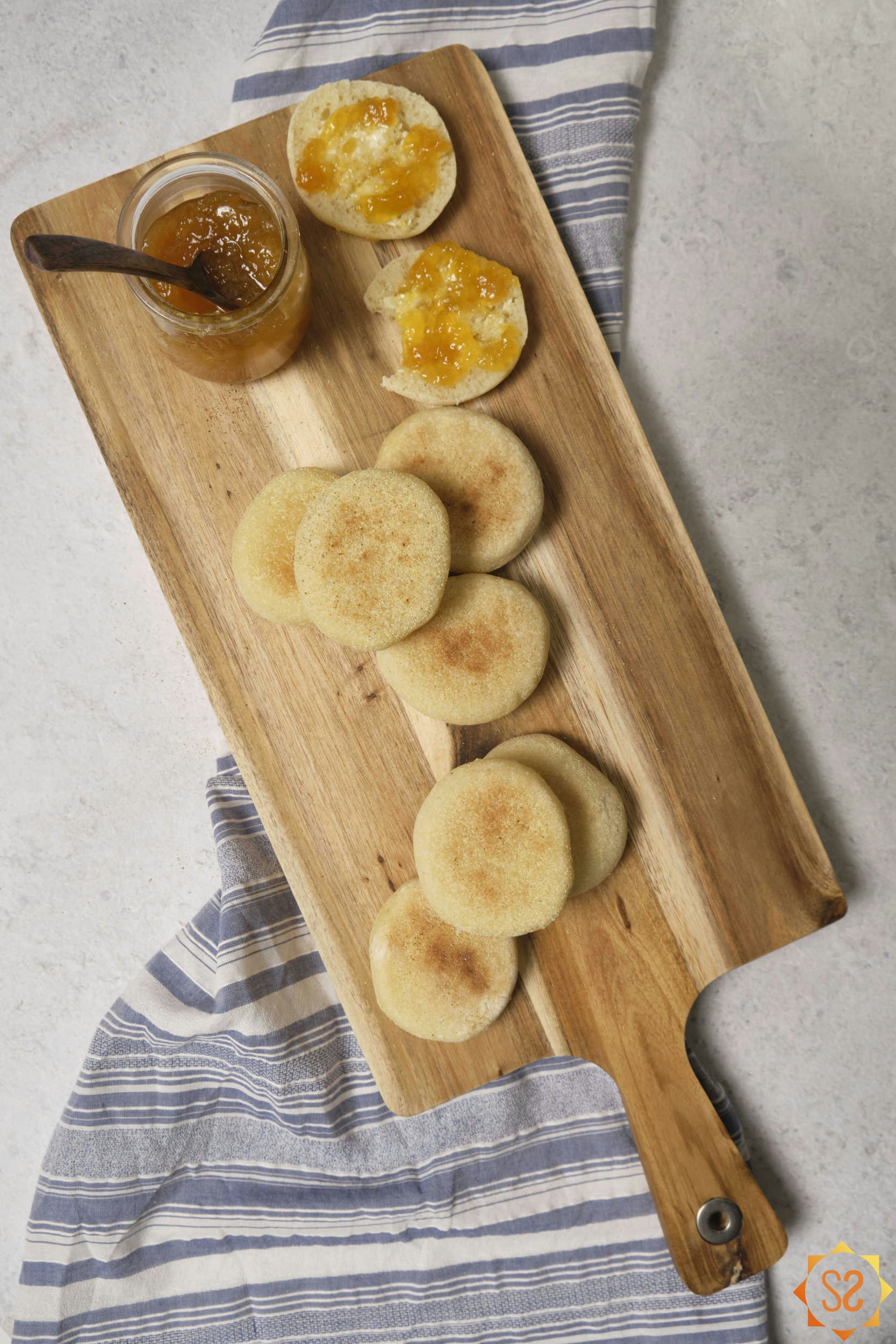 A top down view of English muffins on a serving board, with one at the top sliced open and topped with jelly.