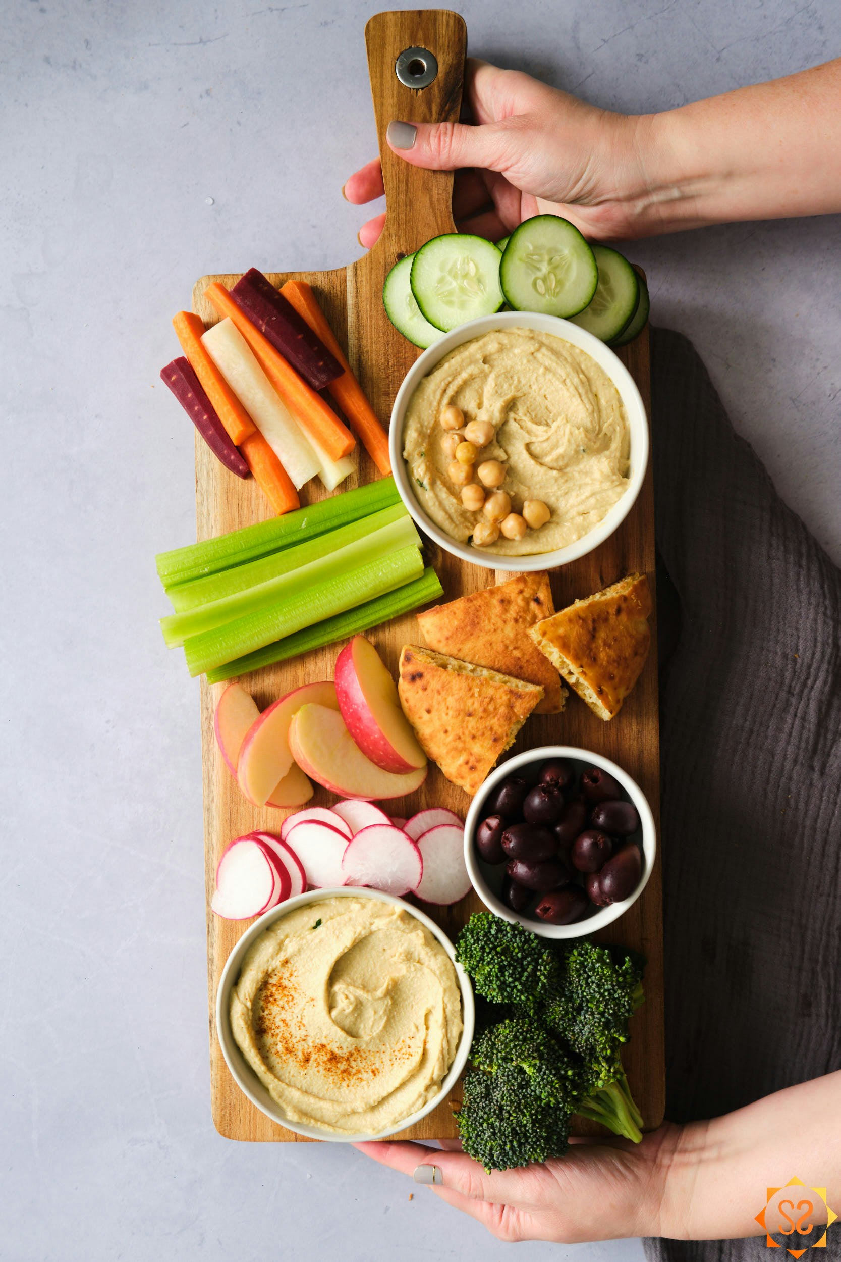 Two hands placing a hummus platter on a table, with two bowls of hummus, olives, vegetables, and pita triangles