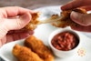 two hands pulling apart a mozzarella stick with vegan cheese stretching in the middle, with sauce and two mozzarella sticks on a plate in the background