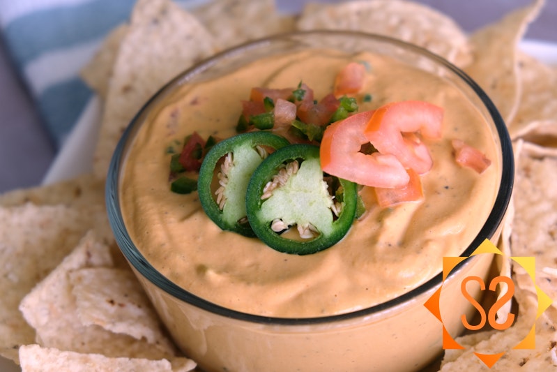 Closeup of vegan queso dip in a bowl topped with jalapeños and tomatoes and surrounded by tortilla chips