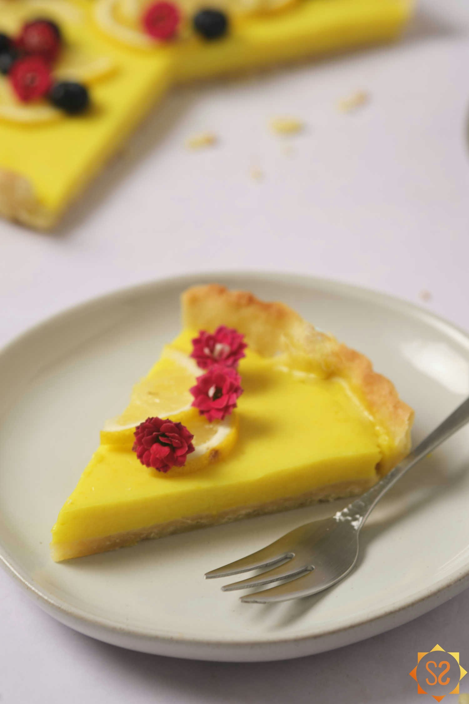 A piece of vegan lemon tart on a plate topped with three flowers; with the rest of the lemon tart in the background.