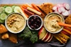 A platter with two bowls of hummus, a bowl of olives, and various vegetables and pita triangles
