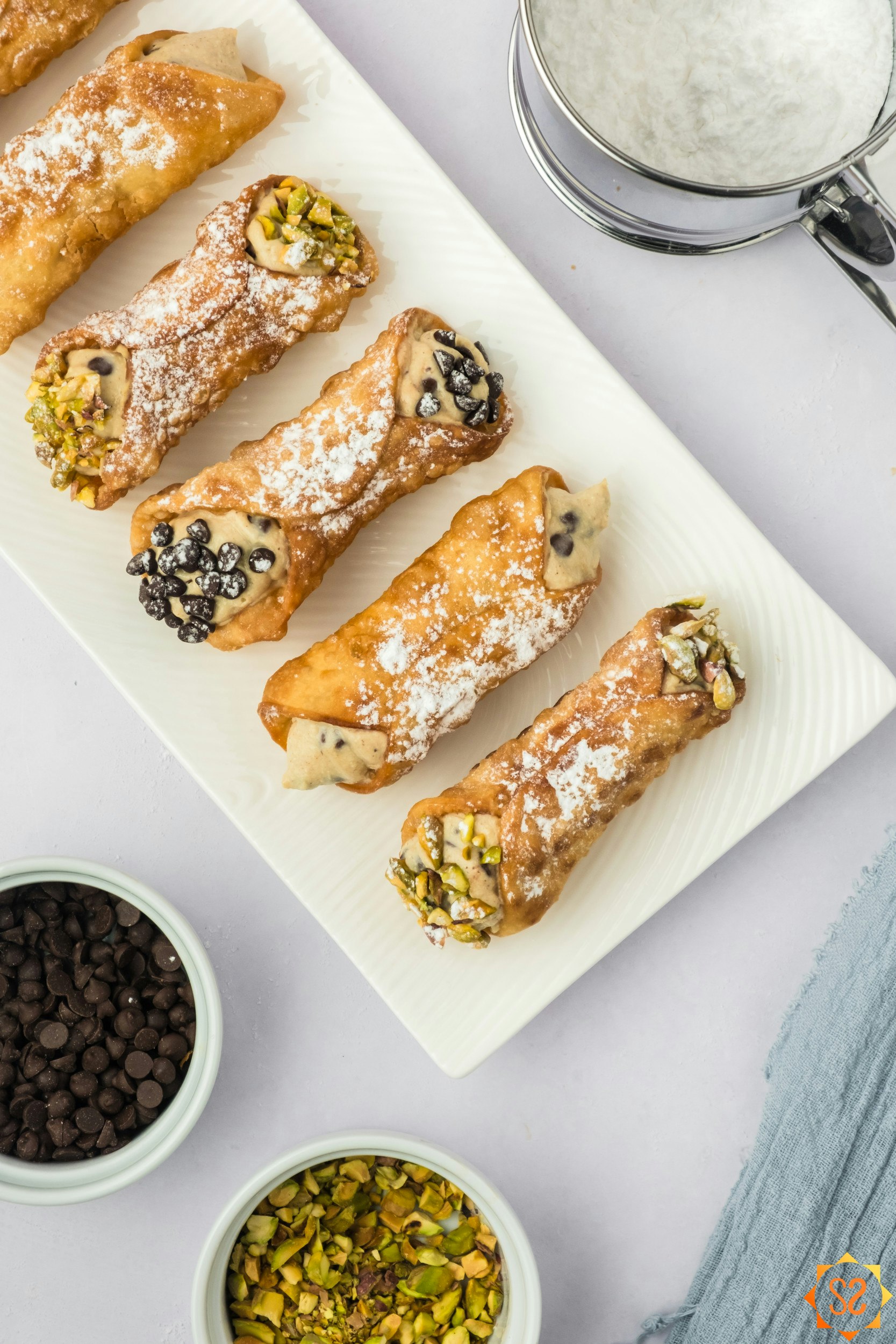 A top-down view of vegan cannoli on a platter with powdered sugar, chocolate chips, and pistachios to the side.