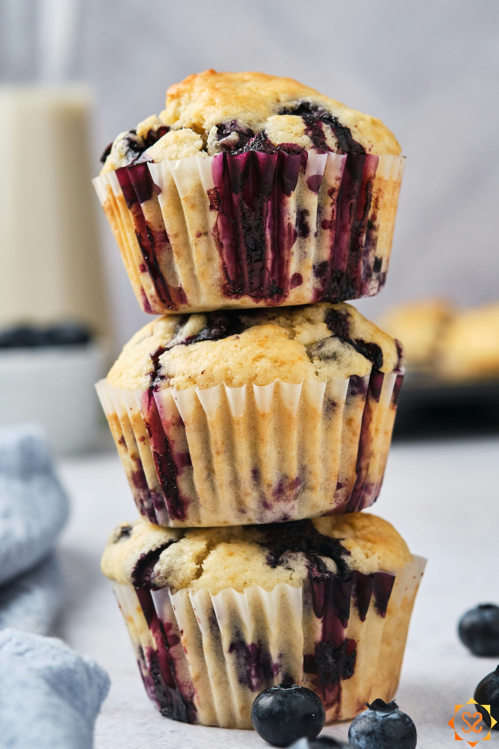 Three vegan blueberry muffins stacked on top of each other, with almond milk and more muffins in the background.
