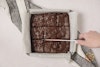 Brownies in a pan with a hand using a knife to slice them.