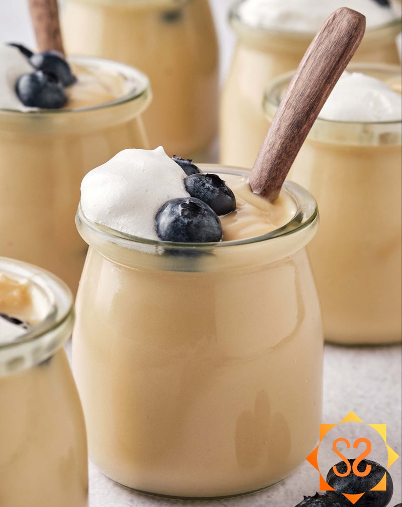 Many jars of vegan vanilla pudding, topped with blueberries and whipped cream, with wooden spoons.