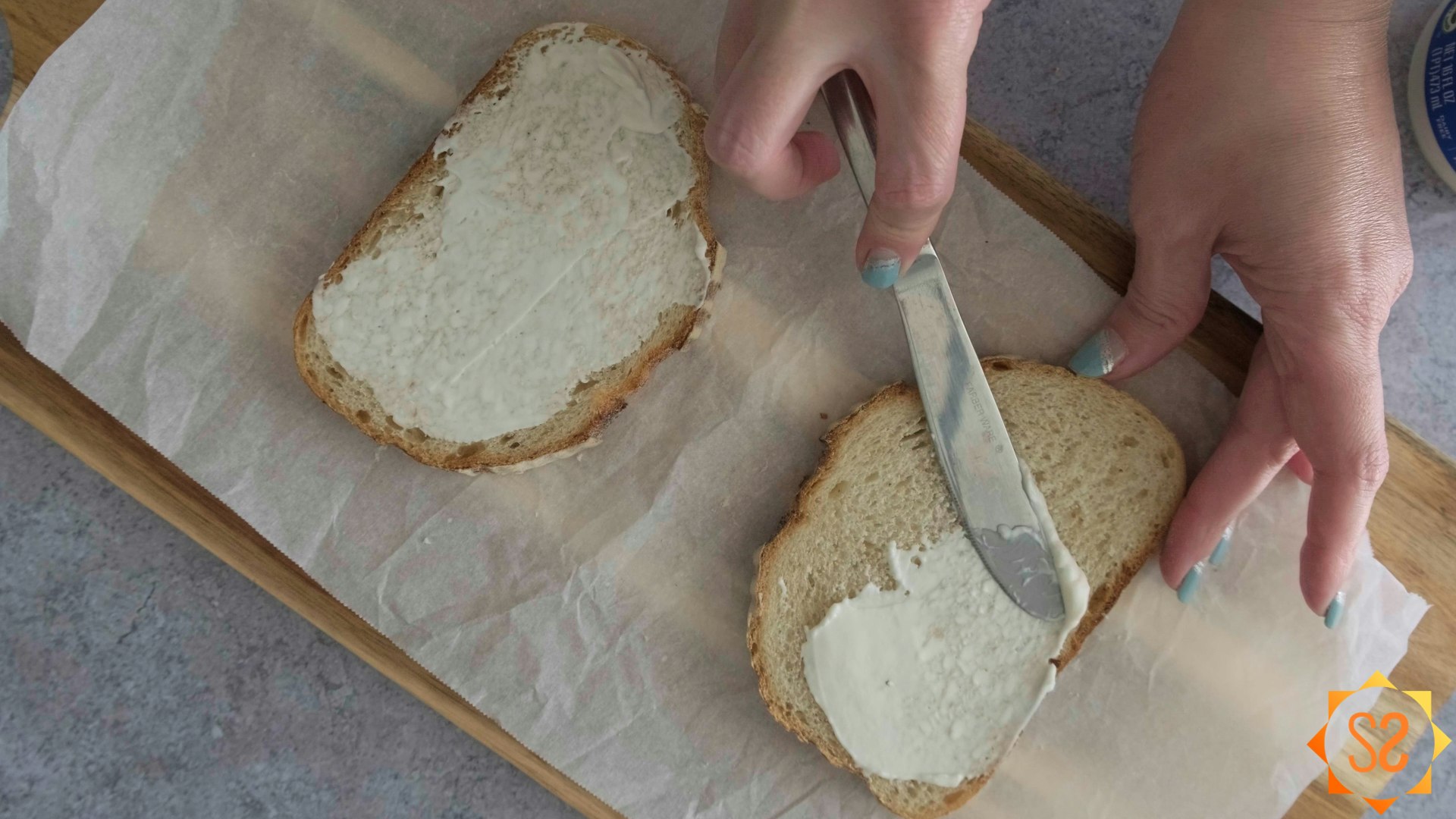A hand spreading vegan mayonnaise on two slices of bread with a butter knife, on top of parchment paper and a serving board