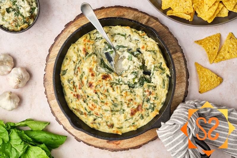 A top-down view of vegan spinach and artichoke dip in a cast-iron pan, with a spoon. To the sides are garlic, spinach, chips, and a bowl of dip.