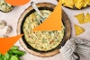 A top-down view of vegan spinach and artichoke dip in a cast-iron pan, with a spoon. To the sides are garlic, spinach, chips, and a bowl of dip.