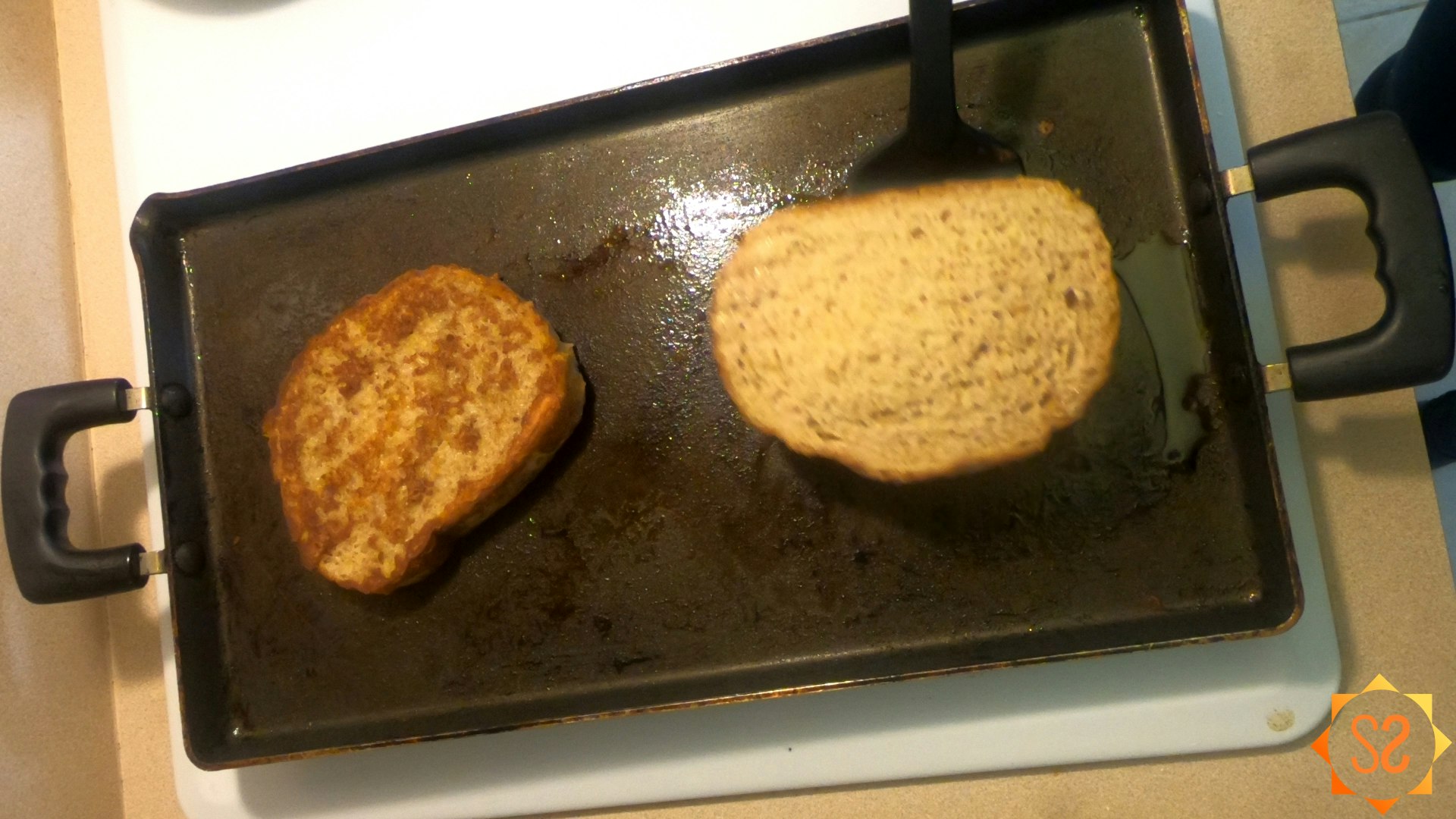 French toast cooking on a griddle, one piece already flipped and the second in the process of being flipped.
