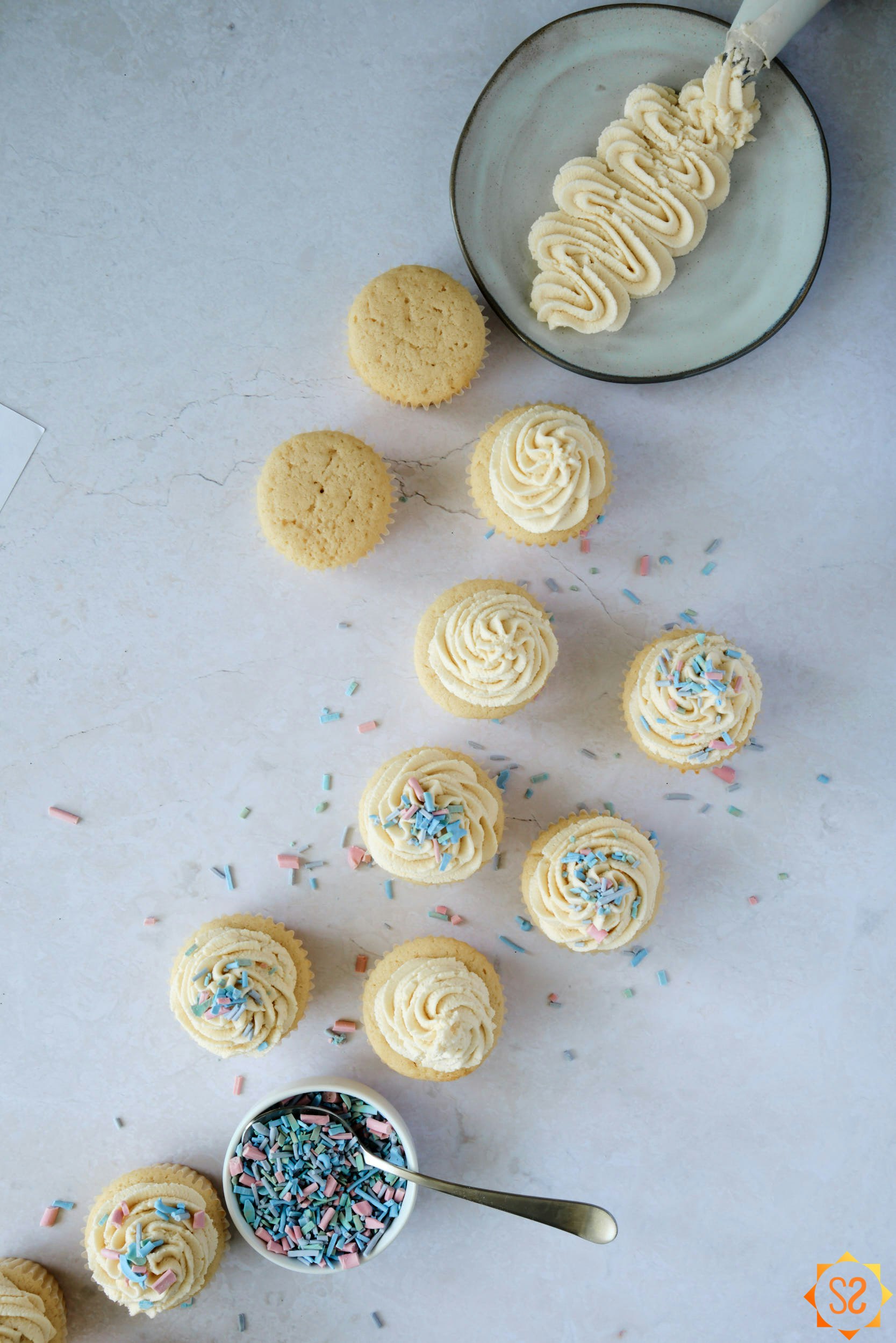 A top-down view of vanilla cupcakes with vegan buttercream frosting, with sprinkles at the bottom, and a plate of buttercream frosting at the top