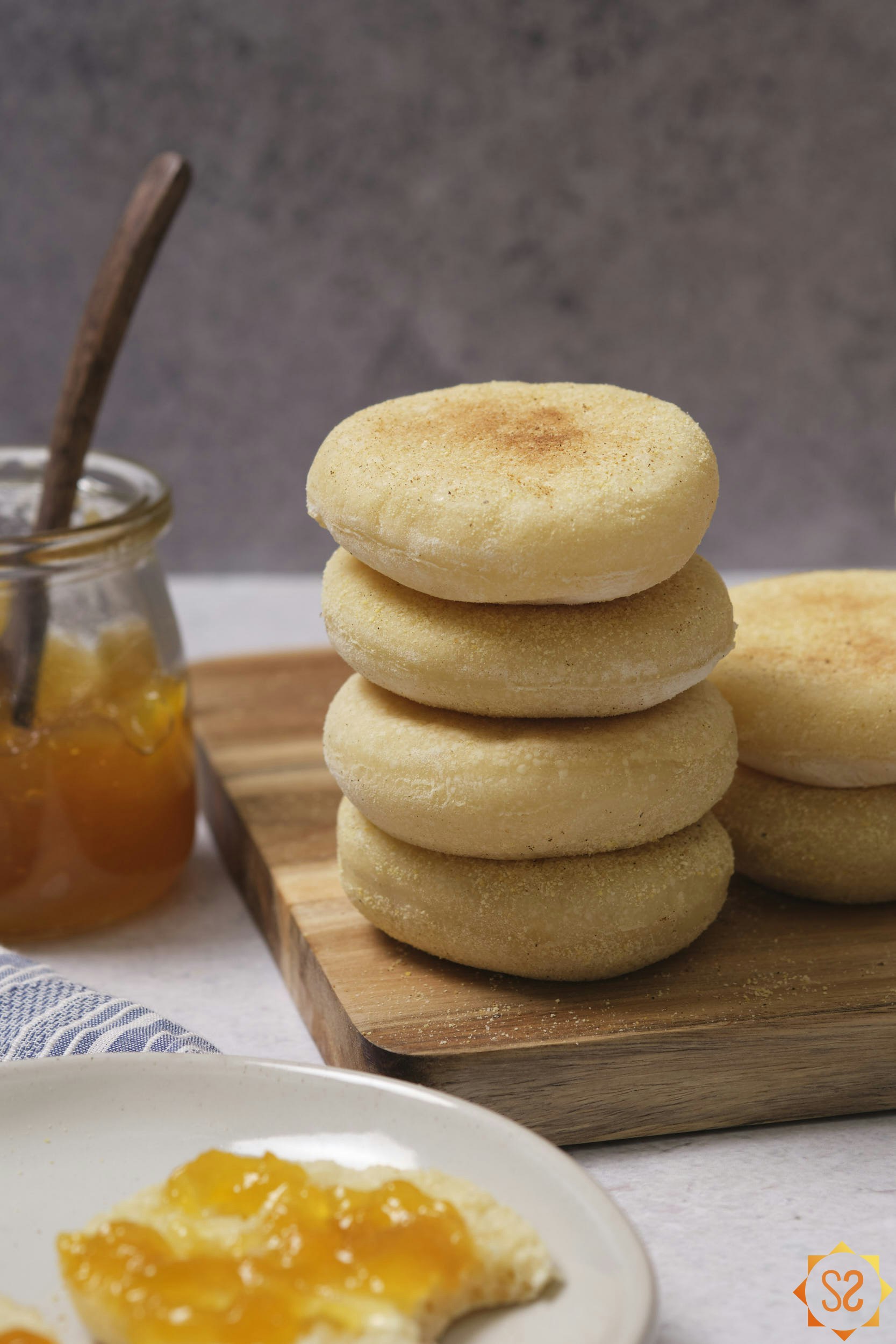A stack of English muffins on a serving board, with a jar of jelly to the side and a cut English muffin topped with jelly to the front.