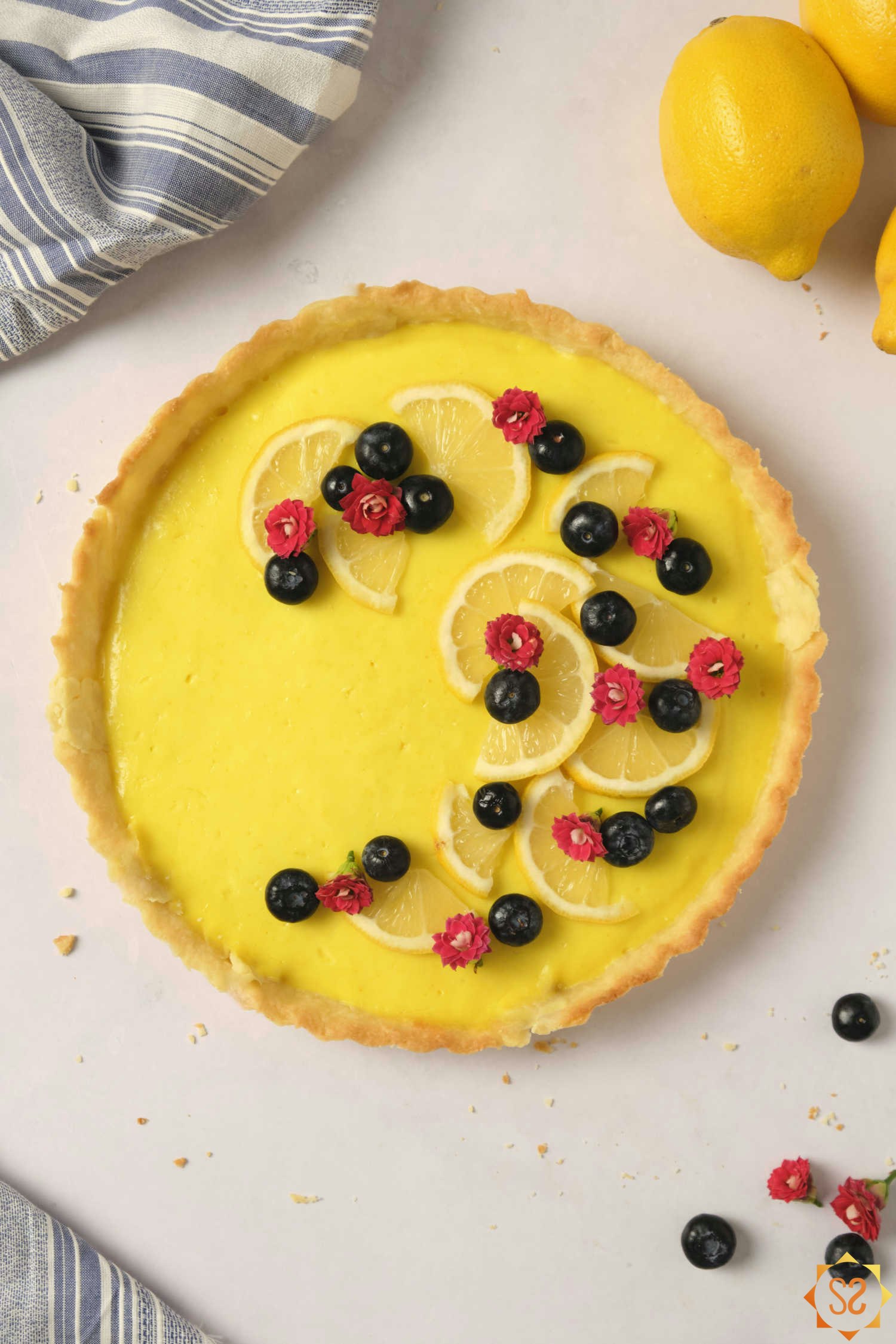 A top-down view of a vegan lemon tart topped with flowers and blueberries, with lemons and a kitchen towel to the side.