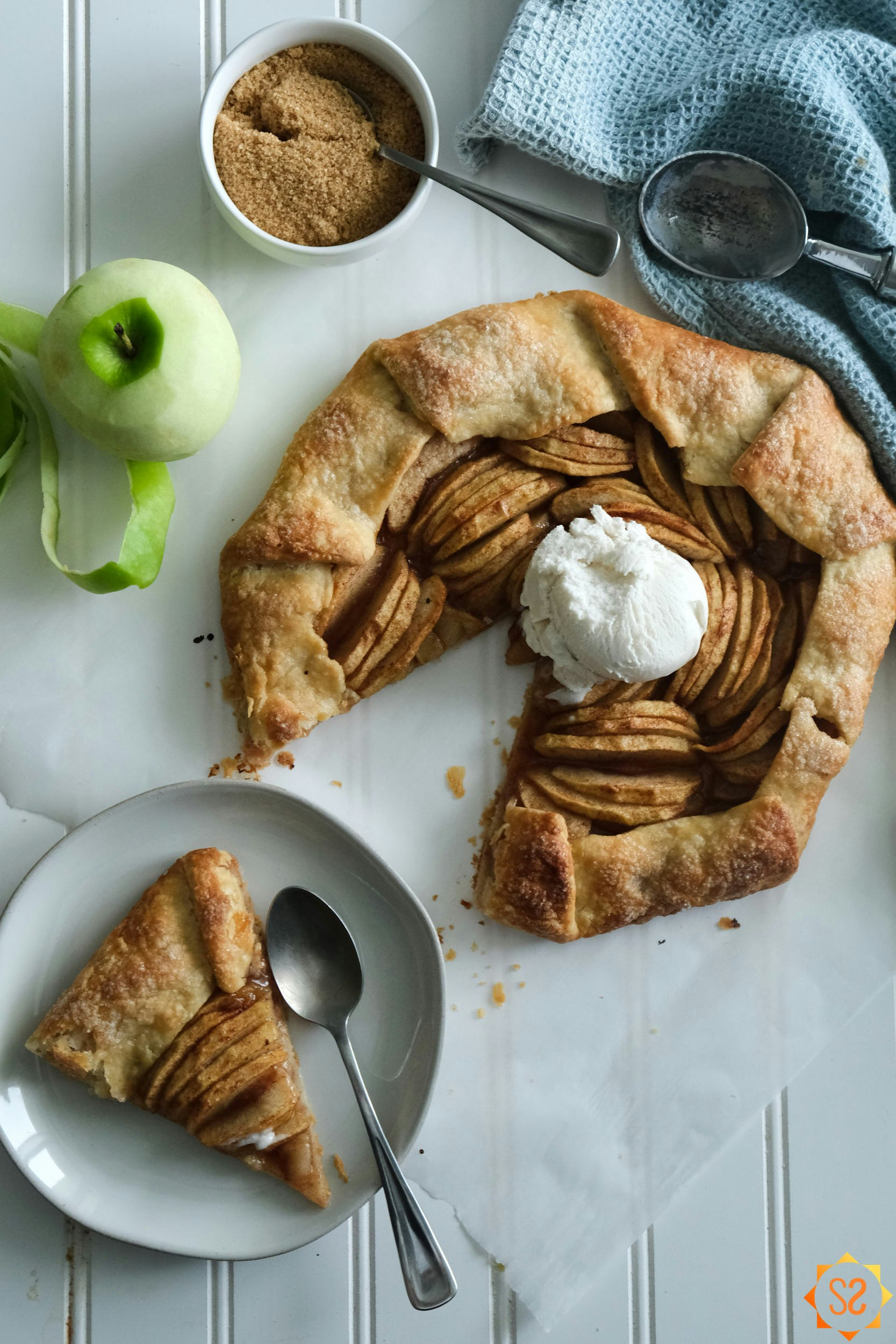A top-down view of an apple galette topped with ice cream, with a slice on a plate, a peeled apple to the side, a ramekin of brown sugar and an ice cream scoop above.