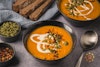 Two bowls of carrot pumpkin soup on a table with bread and toppings.