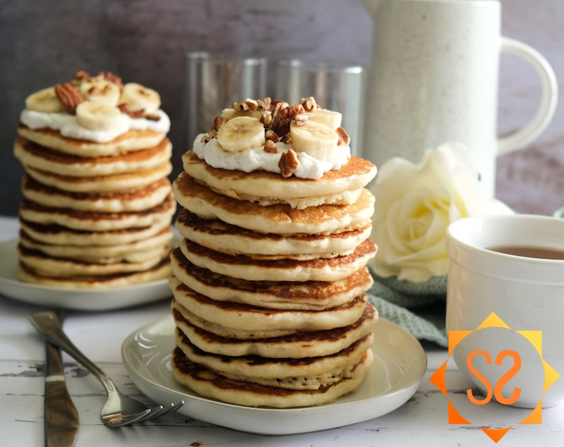 Two stacks of vegan buttermilk pancakes, topped with whipped cream, bananas, and pecans; with a cup of tea, and pitcher of juice