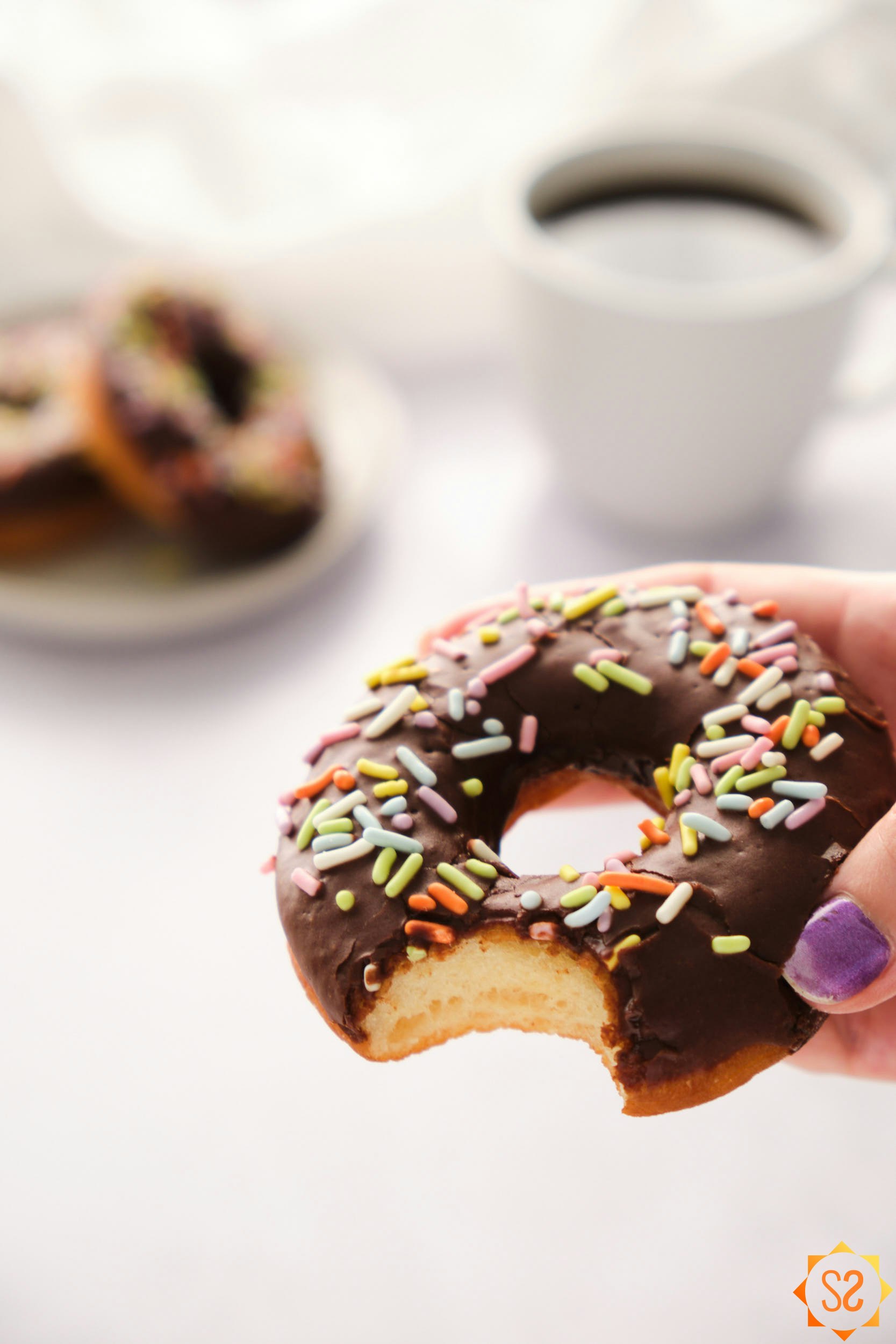 A hand holding a bitten chocolate frosted donut, with a plate of donuts and coffee in the background