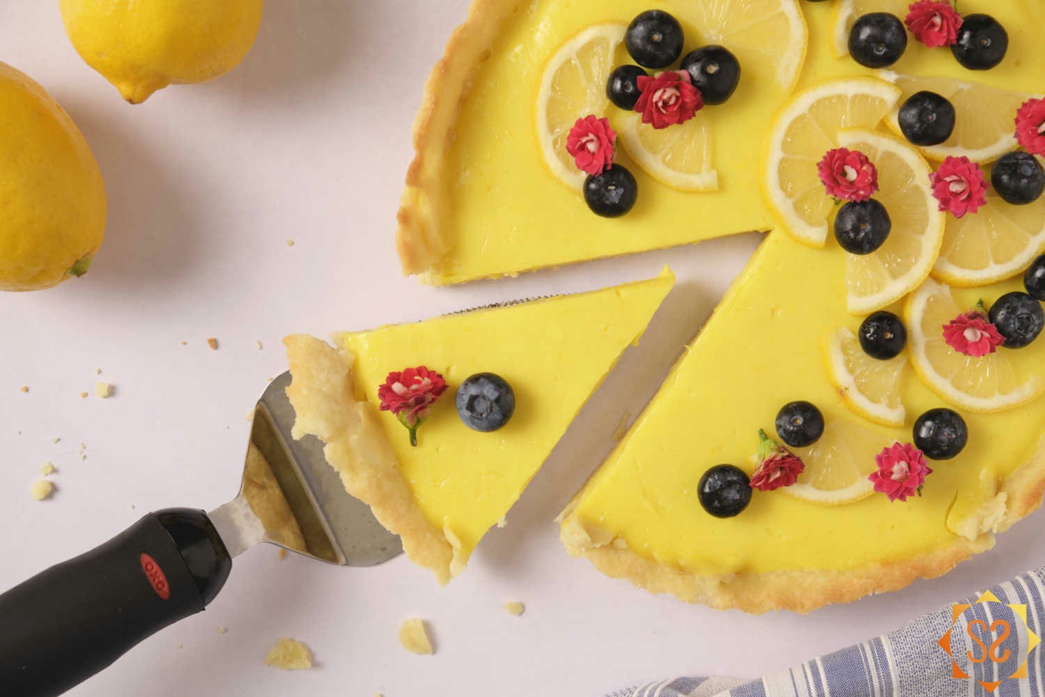 A close-up top-down view of a slice being pulled out from a vegan lemon tart topped with flowers, blueberries, and lemon slices.