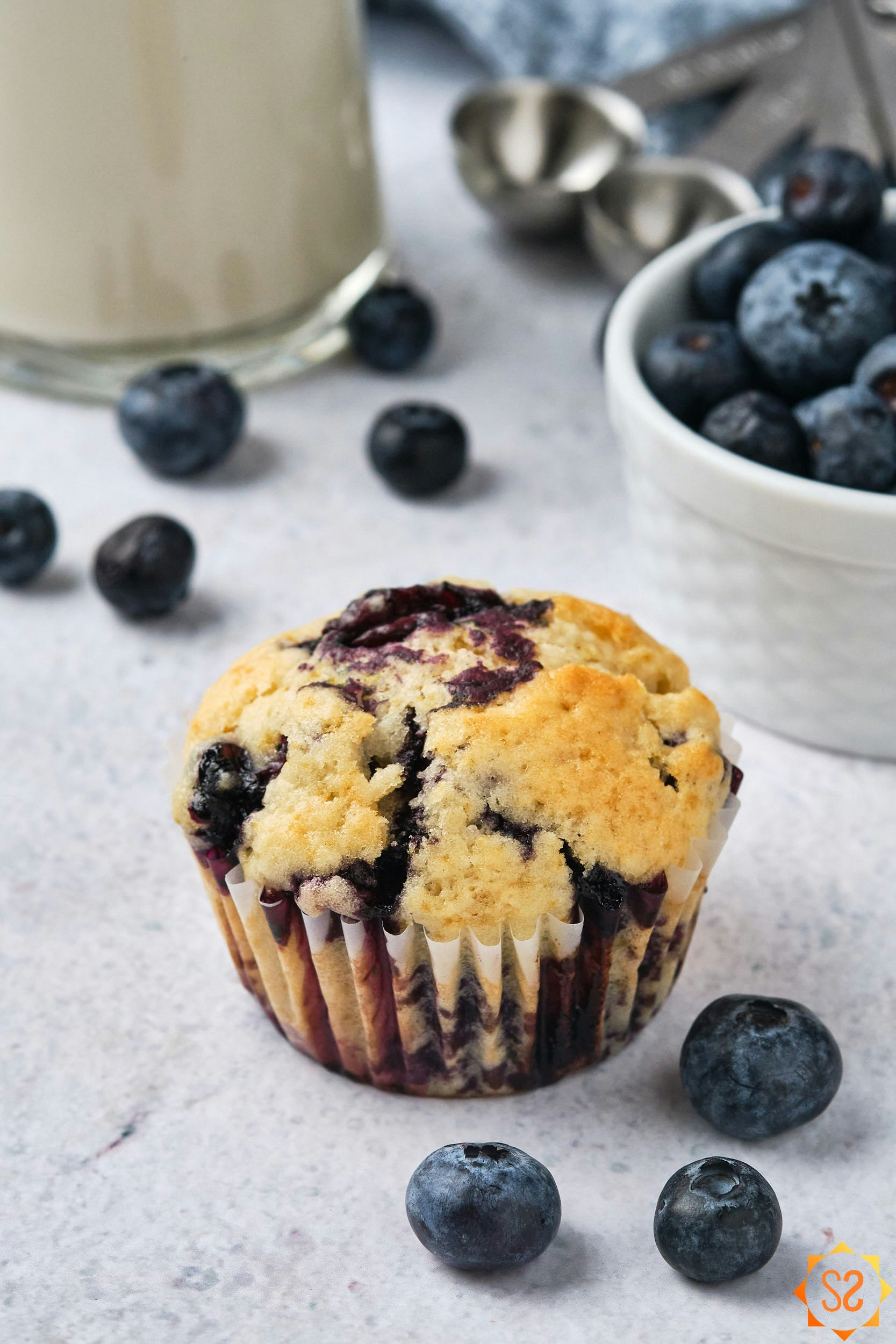 A vegan blueberry muffin, with blueberries, almond milk, and measuring spoons in the background. 