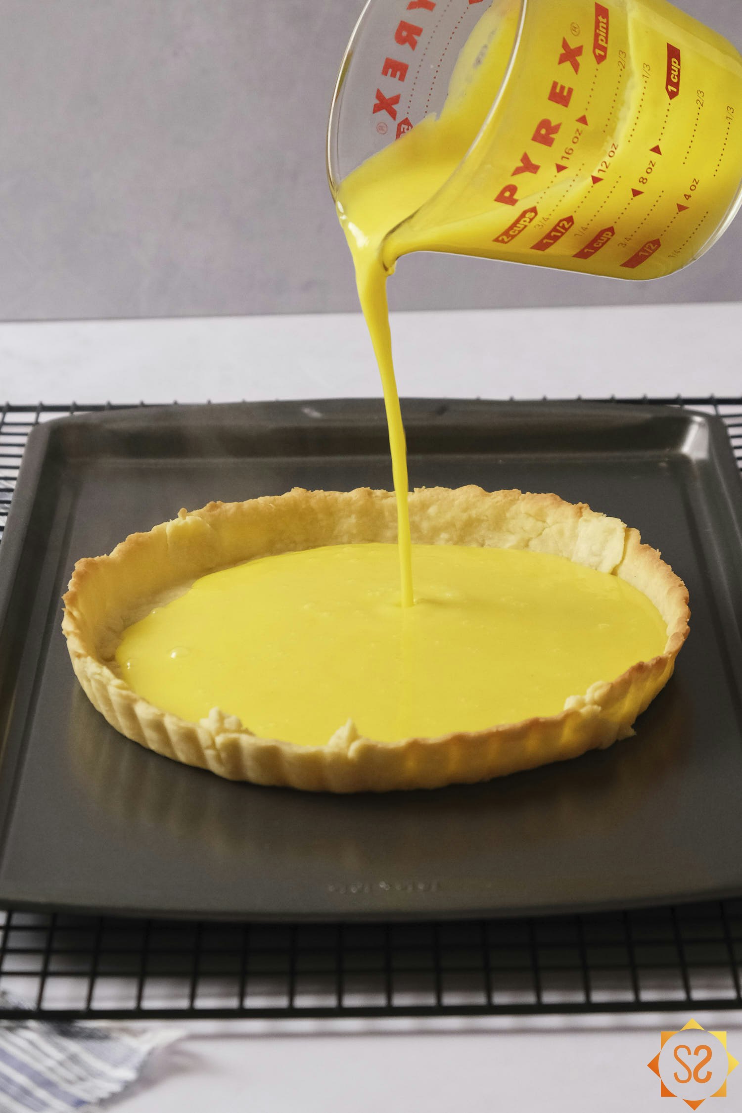 Pouring lemon curd from a measuring cup into the pate brisee crust on a baking tray.