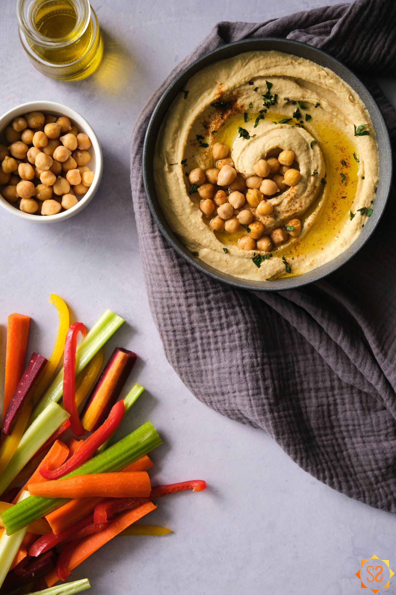 A bowl of hummus with sliced vegetables, garbanzo beans, and olive oil to the side