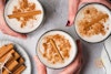 A top-down view of three hands holding three glasses of vegan coquito; a stack of cinnamon sticks is to the side.