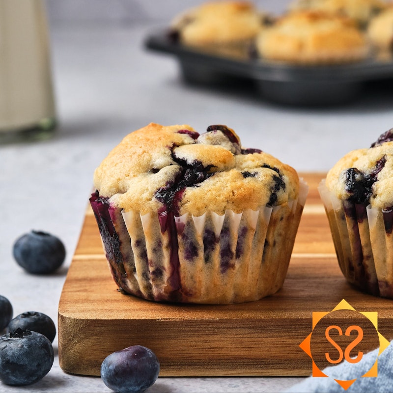 Vegan blueberry muffins on a wooden platter, with plant based milk and a tray of more muffins in the background.