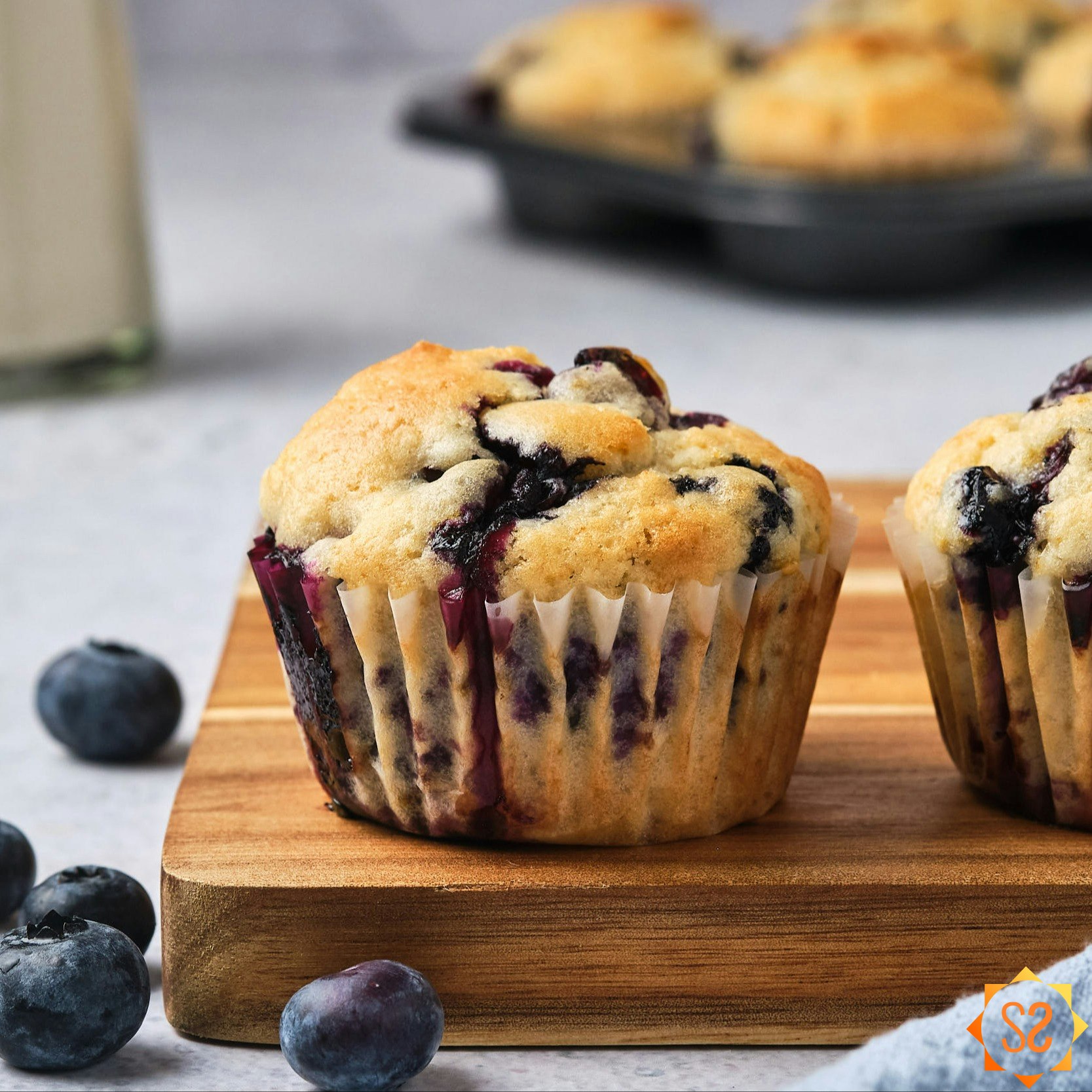 Vegan blueberry muffins on a wooden platter, with plant based milk and a tray of more muffins in the background.