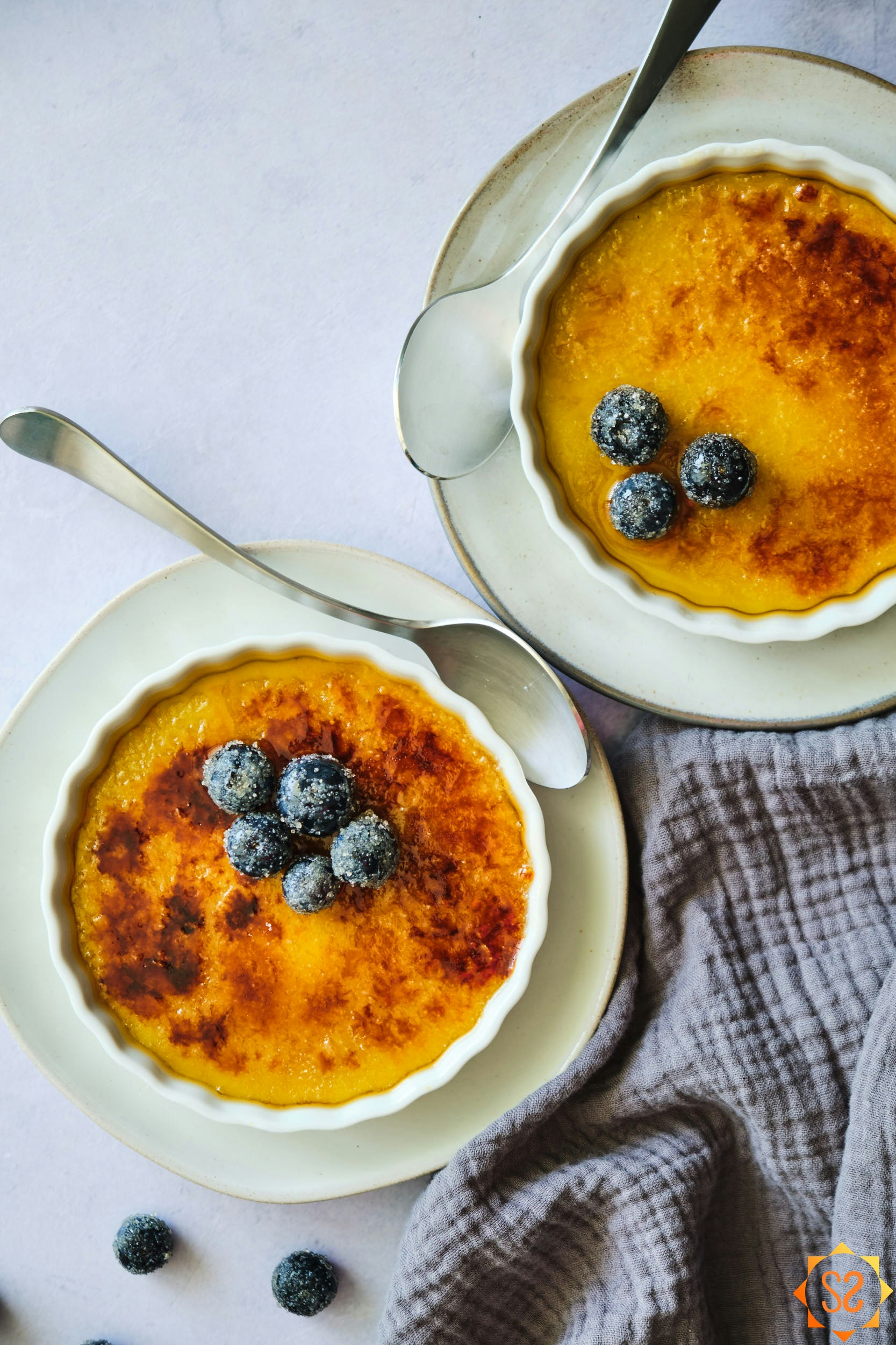 two dishes of vegan creme brulee topped with blueberries, from a top-down angle, with a cloth to the side