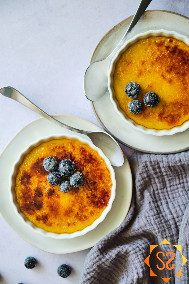 two dishes of vegan creme brulee topped with blueberries, from a top-down angle, with a cloth to the side