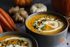 Bowls of carrot pumpkin soup surrounded by carrots, pumpkins, and ginger