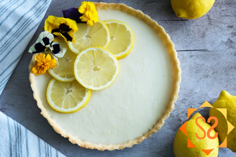 A top-down view of a lemon tart topped with lemon slices and flowers; a kitchen towel and lemons to the sides.