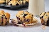 Three vegan blueberry muffins in a row, with the middle one unwrapped; behind them are a pan of muffins and a jug of almond milk.