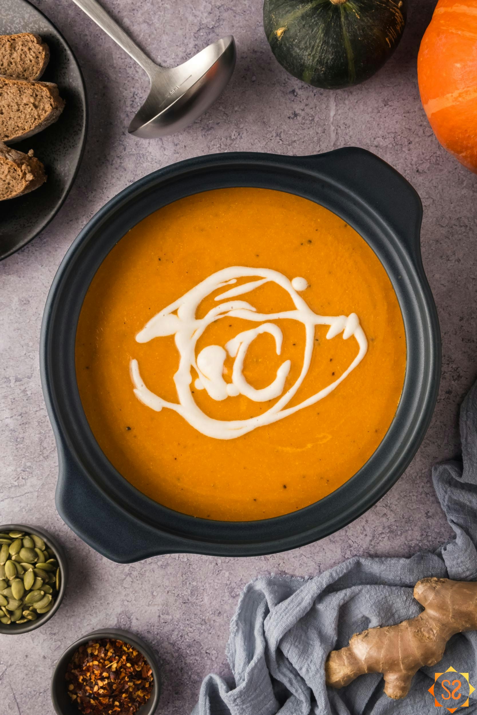 A top-down view of carrot pumpkin soup in a soup pot topped with cashew cream. Surrounding the pot are bread, a ladle, pumpkins, ginger, and bowls of red pepper and pepitas.