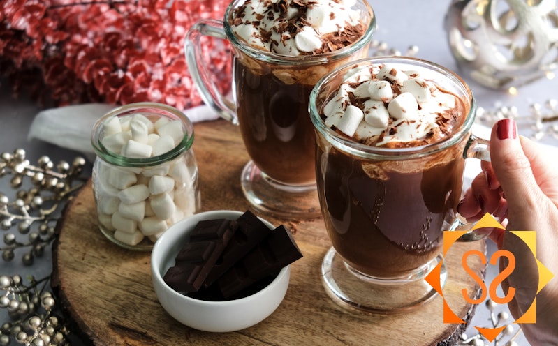 A hand holding one of two mugs of vegan hot chocolate, next to a jar of marshmallows and a dish of chocolate pieces