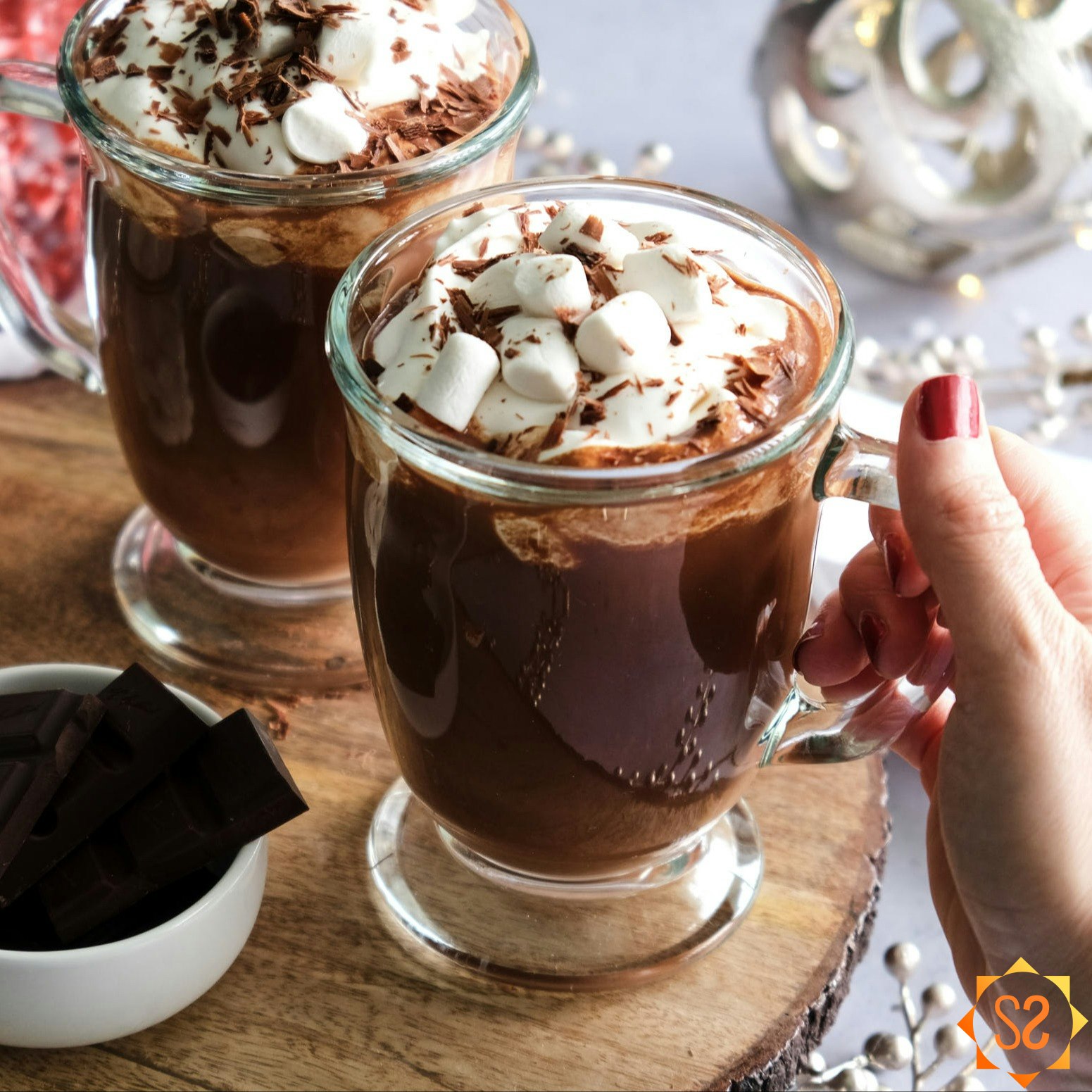 A hand holding one of two mugs of vegan hot chocolate, next to a jar of marshmallows and a dish of chocolate pieces.
