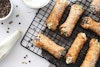 vegan cannoli on a cooling rack with powdered sugar, chocolate chips, and a piping bag of filling on the side.