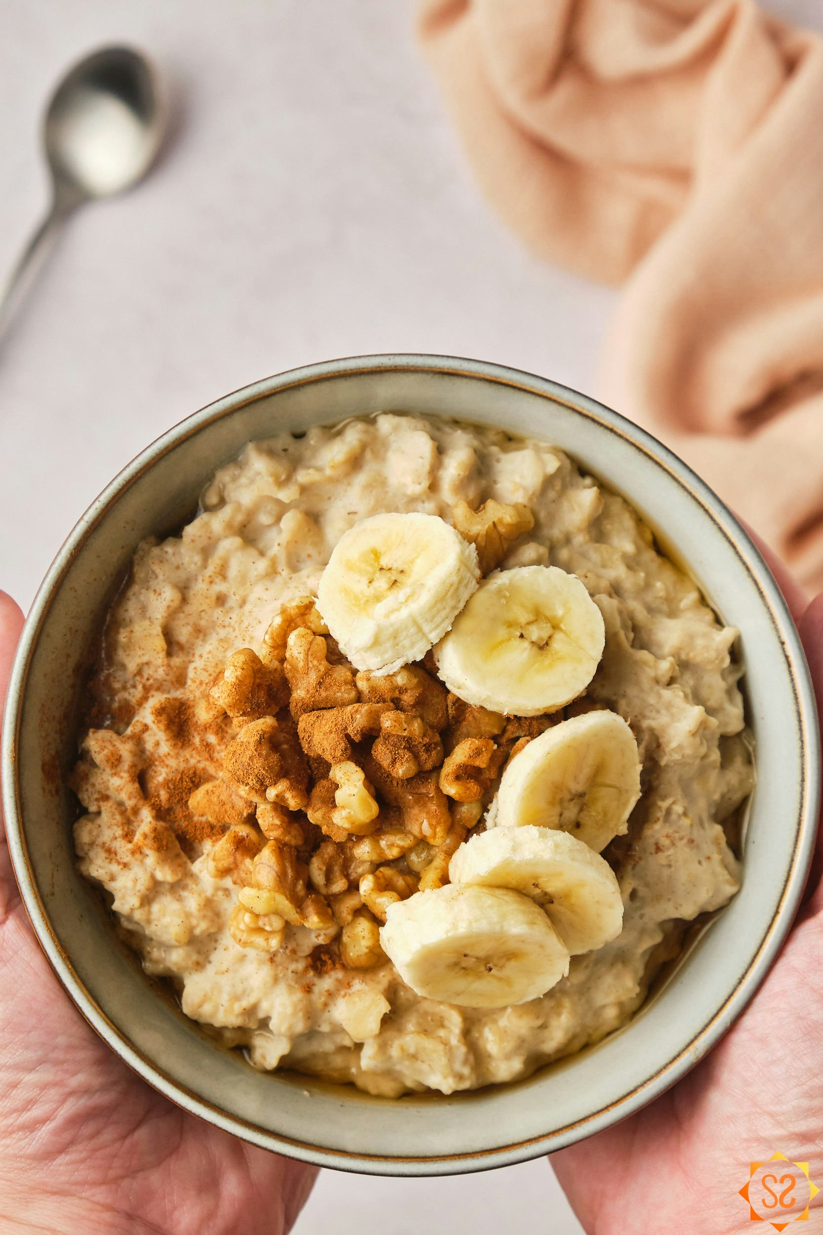 Two hands holding a bowl of protein oatmeal for a close view, topped with banana slices, peanut butter, and walnuts