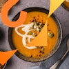 A top-down image of a bowl of creamy carrot pumpkin soup topped with cashew cream, red pepper flakes, and pepitas.