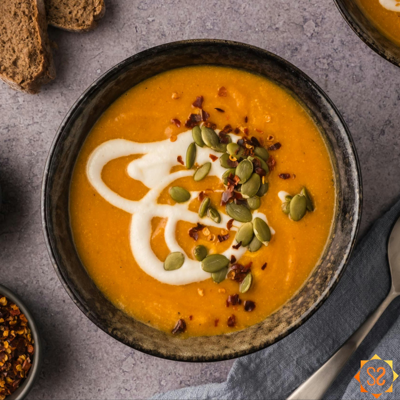 A top-down image of a bowl of creamy carrot pumpkin soup topped with cashew cream, red pepper flakes, and pepitas.